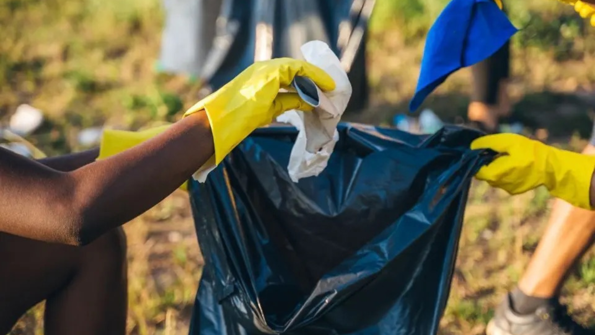 A group of hands surrounds a trash bag. One person is putting a piece of trash into the garbage bag as part of a community clean up project.