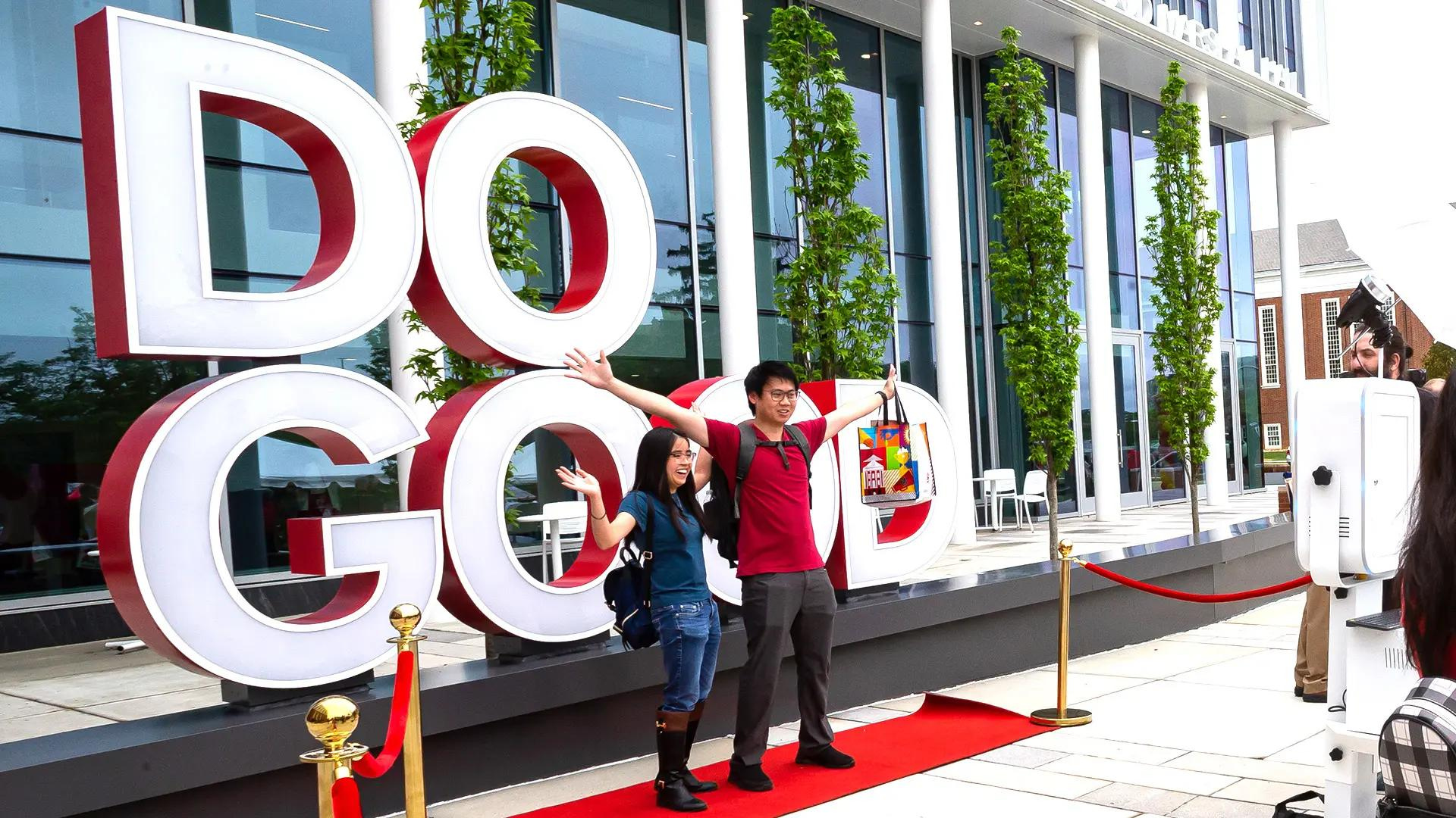 A man and a woman are standing with arms raised in front of a large Do Good sign.
