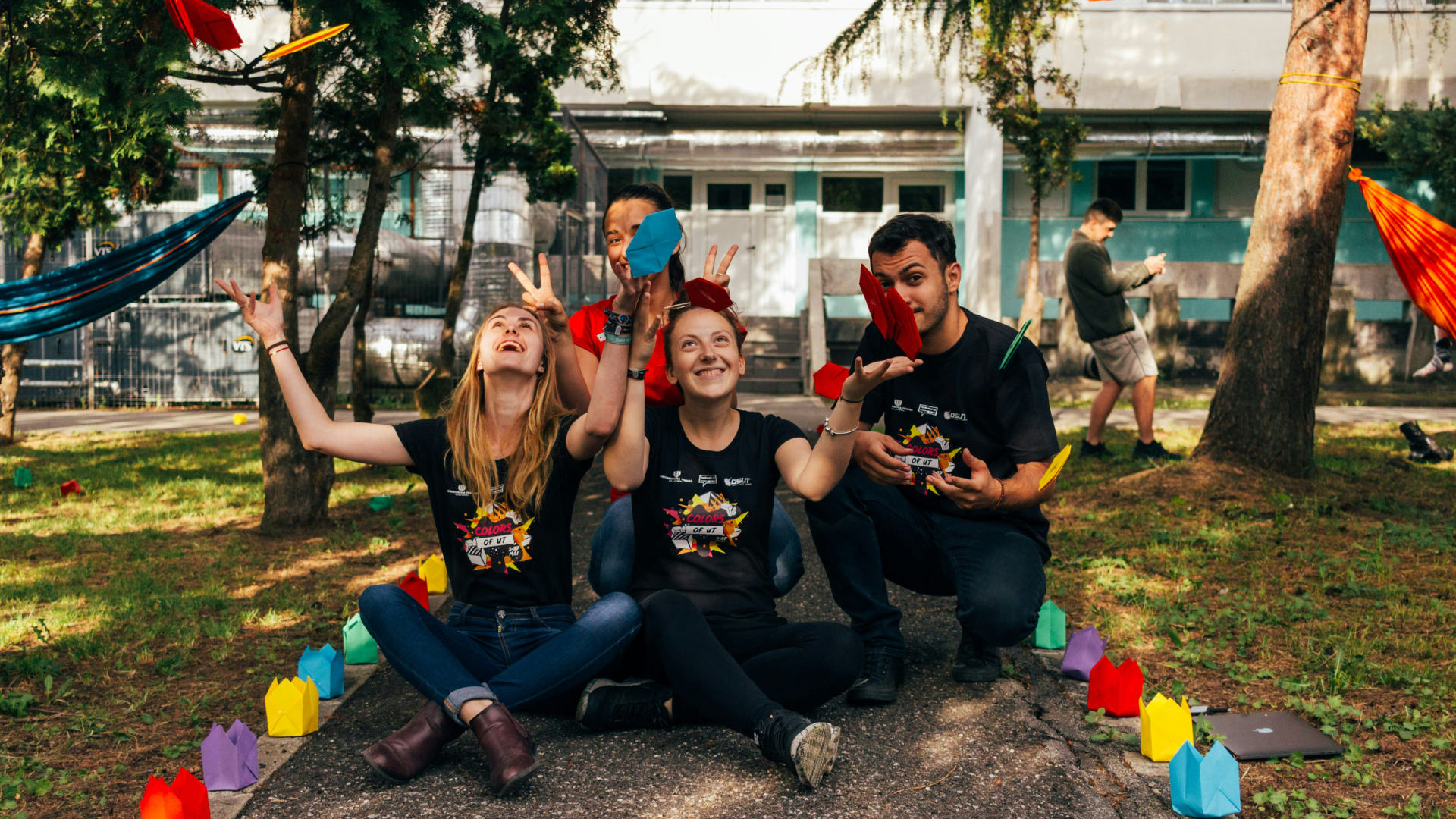 A group of three volunteers sitting down cross legged. Wearing black tshirts, they are throwing colorful pieces of fabric in the air.