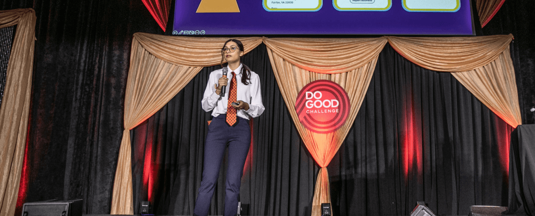 Kat Close holding a mic on stage at the Do Good Challenge with gold drapes in the background