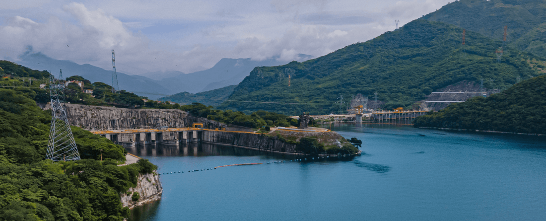 An aerial view of a hydropower generation station with bright blue water in the foreground surrounded by forest-covered mountains.