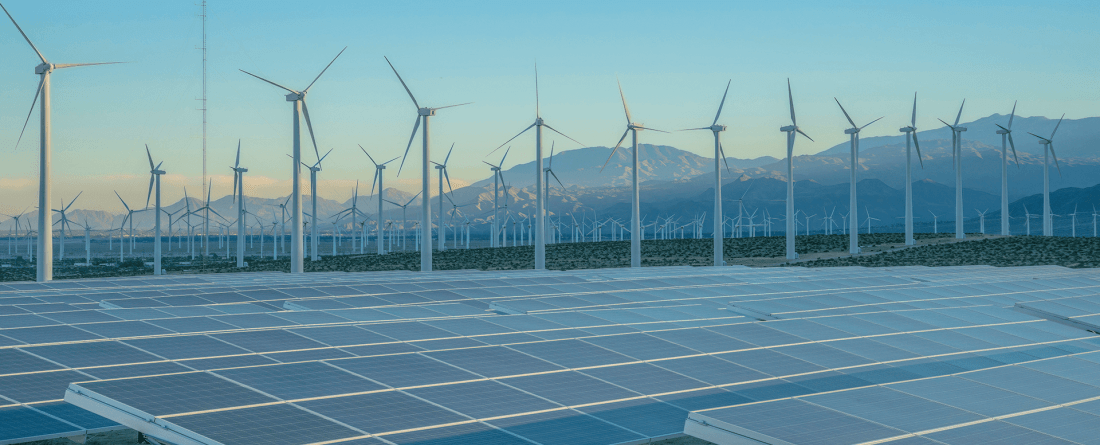 A field of solar panels and wind turbines in a valley area surrounded by mountains during blue hour.