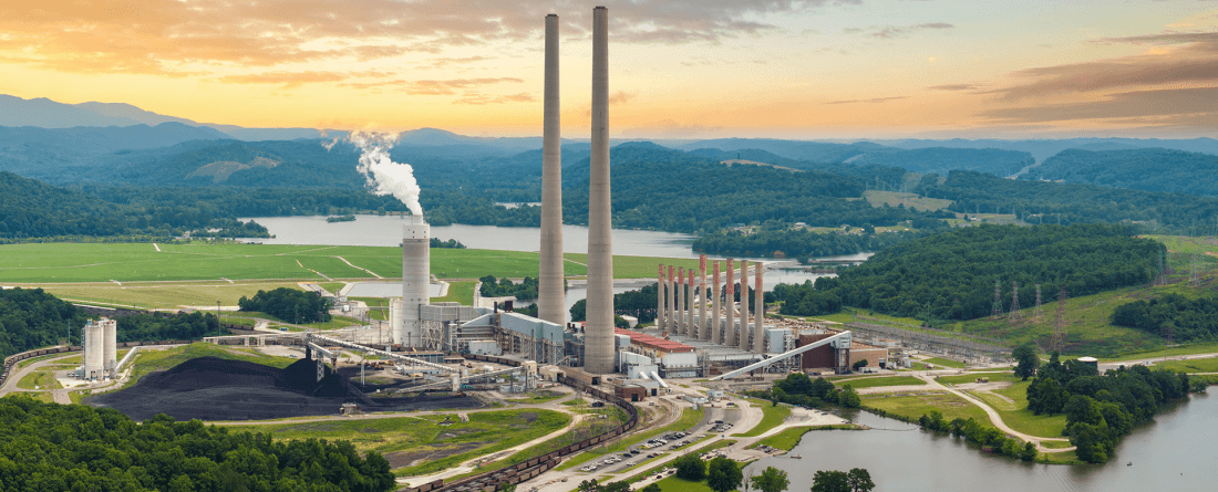 A large powerplant with small smokestack on the bank of a river with green grass. The sky is blue and orange with some clouds.