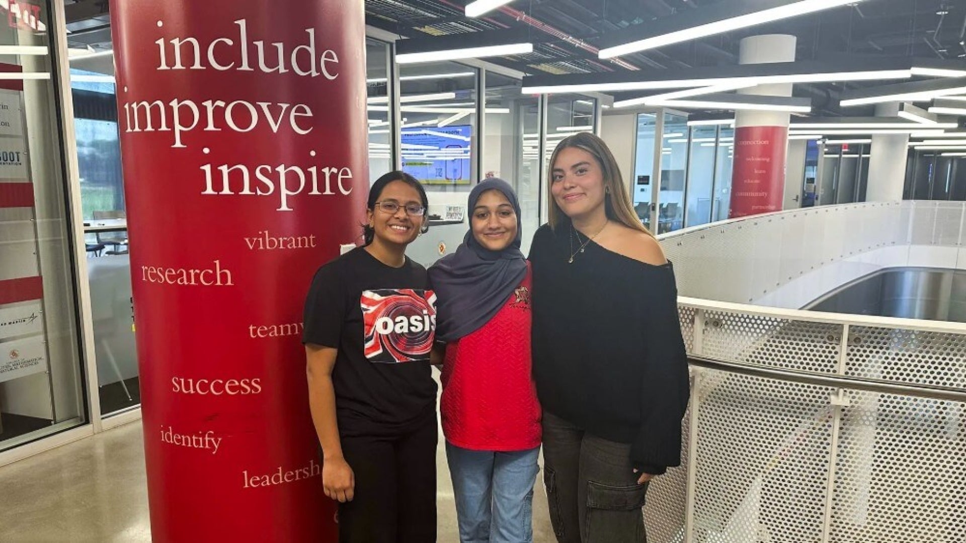 2 female students standing next to a red wrapped pole that says "include, improve, inspire" in big white letters, then in smaller white letters it says "vibrant, research, team, success, identify, leadership"
